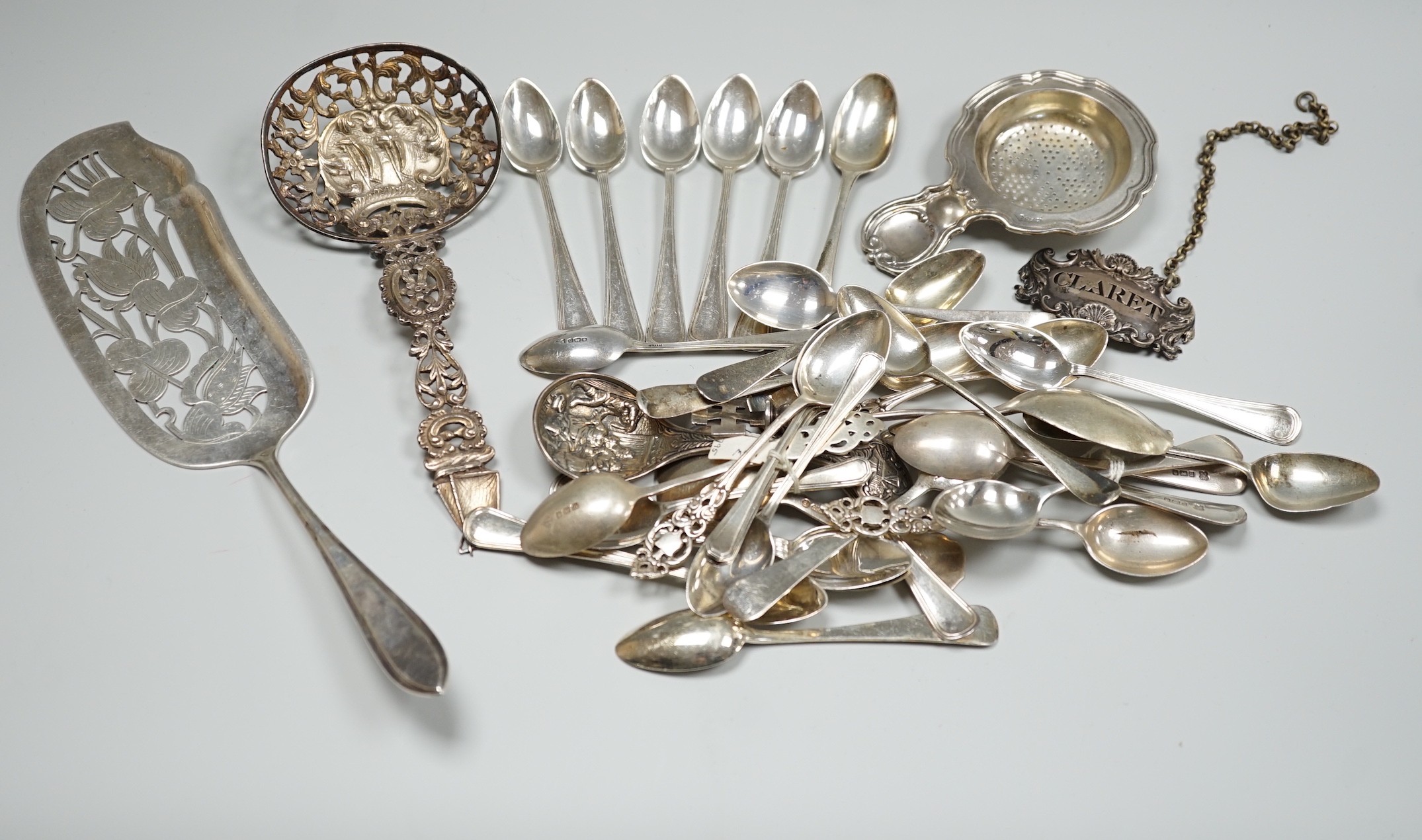 A group of mixed silver and white metal items including flatware, Dutch fish slice, German 800 tea strainer, George IV 'Claret' wine label, etc. and a plated spoon.